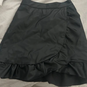 Svart omlottkjol med volang - Snygg svart omlottkjol med volangkant nertill. Kjolen har en stilren design och passar perfekt till många olika outfits. Den har dessutom ett skinnmaterial. 💗