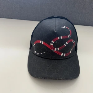 Svart keps med ormtryck från Gucci - Snygg svart keps från Gucci med ett detaljerat rött, vitt och svart ormtryck framtill. Kepsen har mesh baktill och justerbar rem med röd och blå detalj. Perfekt för dig som vill sticka ut med en exklusiv accessoar.