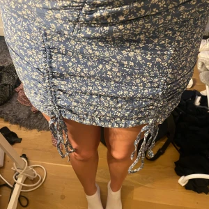Blå blommig kortkjol med snörning - Säljer en blå kortkjol med vitt blommönster och snörningsdetaljer på båda sidor. Kjolen har dragkedja på sidan och är stretchig. 😍 Den är liten på mig som har 36 i byxor:)