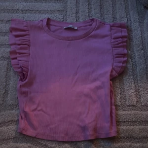 Rosa ribbad topp med volangärmar - Supersöt rosa topp med ribbad struktur och volangdetaljer på ärmarna. Perfekt för dig som vill ha en feminin och trendig look. Rund halsringning och kort ärm med volang ger en extra touch.