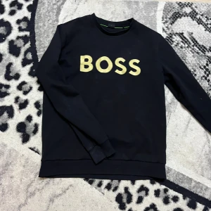 Svart sweatshirt från BOSS - Säljer en svart sweatshirt från BOSS med stor gul logotyp på bröstet. Tröjan har rund halsringning och långa ärmar. Perfekt för en stilren och avslappnad look.