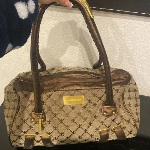 Brun mönstrad handväska från Friis & Company - Säljer en brun handväska från Friis & Company med klassiskt mönster och detaljer i guld. Väskan har två rejäla handtag, dragkedja upptill och flera guldfärgade detaljer. Perfekt för dig som vill ha en snygg accessoar med plats för det mesta.