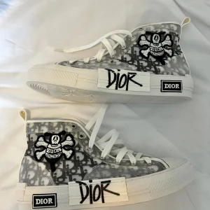 Dior B23 high-top X Stussy - Säljer ett par Dior B23 high-top sneakers i vitt och grått med tydligt Dior-mönster och graffiti-detaljer på sidan i collab med Stussy. Skorna har vita snören, platt sula och rund tå. Perfekta för dig som vill sticka ut med exklusiva och trendiga sneakers. Storlek 44 men liten i storlek så passar mer som en 43. Har både lådan och påse. Lådan är dock i dåligt skick.