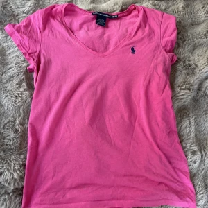Rosa t-shirt från Ralph Lauren Sport - Säljer en rosa t-shirt från Ralph Lauren Sport i 100% pima bomull. T-shirten har v-ringning, korta ärmar och en broderad marinblå logga på bröstet. Perfekt för sommaren🥰! Den är äkta, har aldrig används heller. Kontakta mig privat för fler bilder!💓💓