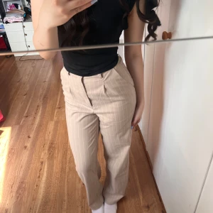 Beige kostymbyxor med vita ränder från H&M Divided - Säljer ett par beige kostymbyxor från H&M Divided med vita tunna ränder. Byxorna har normal passform, raka ben och hög midja. Perfekta för en stilren look och passar till många olika outfits.
