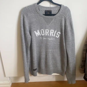 Grå stickad tröja från Morris - Säljer en grå stickad tröja från Morris Company med vit broderad logga framtill. Tröjan har rund halsringning och långa ärmar med ribbade muddar vid ärmslut och nederkant. Perfekt för lager-på-lager under kyliga dagar.