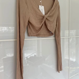 Beige croppad topp med knyt  - Trendig beige croppad topp  med lång ärm och snygg knytning framtill. Toppen har en v-ringad design och är tillverkad i ett ribbat material som ger en cool och modern look.