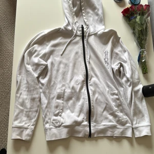 Vit hoodie med svart dragkedja och tryck - Säljer en vit hoodie från Hugo med svart dragkedja och tryck i svart på både bröstet och ryggen. Tröjan har huva med dragsko och två fickor framtill. Perfekt för en avslappnad stil.