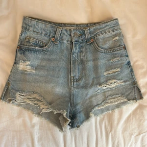 Ljusblåa jeansshorts med slitningar - Säljer ett par ljusblåa jeansshorts med råa kanter och flera slitningar framtill. Klassisk femficksmodell med hög midja och knappgylf. Perfekta för sommaren!