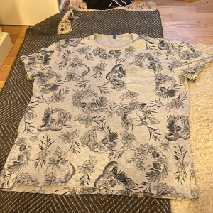 Grå t-shirt med dödskallemönster från H&M Divided - Säljer en grå t-shirt från H&M Divided med svart tryck av dödskallar, blommor och blad. T-shirten har rund halsringning och korta ärmar. Perfekt för dig som gillar lite edgy stil.