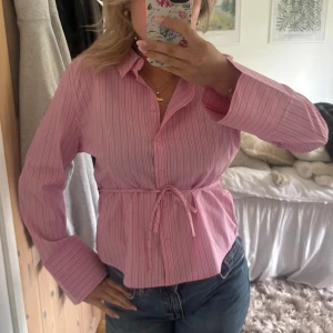 Rosa randig skjorta med knyt i midjan - Säljer en rosa skjorta med tunna vita ränder och knytband i midjan. Skjortan har klassisk krage, knappar framtill och långa ärmar med breda manschetter. Den är aldrig använd med lappar kvar.💕💕🩷