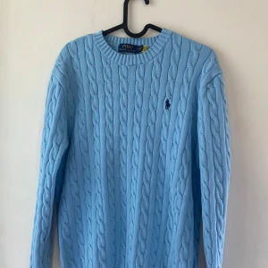 Ljusblå stickad tröja från Polo Ralph Lauren - Säljer en ljusblå kabelstickad tröja från Polo Ralph Lauren med rund halsringning och broderad logga på bröstet. Tröjan har långa ärmar och klassisk passform. Perfekt för lager-på-lager under kyliga dagar.