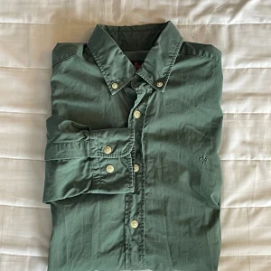 Grön skjorta från Morris - Snygg grön skjorta från Morris med klassisk krage och broderad logga på bröstet. Skjortan har långa ärmar och knäppning framtill med vita knappar. Perfekt för dig som gillar stilrena plagg med diskreta detaljer.