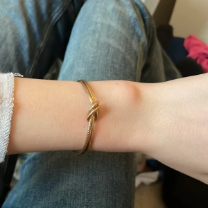 Guldfärgat armband med knutdetalj - Snyggt armband i guldfärg med en dekorativ knut framtill. Armbandet är stelt och har en öppen design som gör det enkelt att ta på och av samt justera storleken. Perfekt accessoar för att lyfta din outfit.