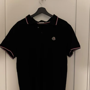Svart pikétröja från Moncler - Säljer en svart pikétröja från Moncler med klassisk krage och knappslå. Tröjan har Moncler-logga på bröstet och diskreta rödvita detaljer på krage och ärmslut. Perfekt för en stilren och sportig look. Passar L & XL