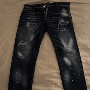 Mörkblå jeans dsquared2 - Säljer ett par mörkblå jeans från Ciao Bella med slitna detaljer, färgstänk och tryckt text på bakfickan. Jeansen har knappar i gylfen och en klassisk femficksdesign. Perfekta för dig som gillar en edgy och unik stil. 