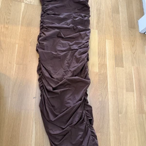 Brun långklänning med rynkade detaljer - Säljer en brun långklänning med snygga rynkade detaljer längs sidorna. Klänningen har en tight passform och är perfekt för dig som gillar en stilren look. Materialet känns mjukt och följsamt mot kroppen. helt ny med prislappen på