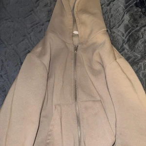 Hoddie - Säljer en beige hoodie med dragkedja framtill och huva. Tröjan har två fickor på magen och är tillverkad i ett mjukt material, perfekt för en avslappnad stil. Använd endast 1 gång pga fel storlek, som nytt skick!!