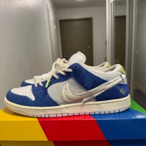 Nike SB Dunk Low Fly Streetwear blå/vit - Nike SB Dunk Low i samarbete med Fly Streetwear. Skorna har blå mockadetaljer, vita läderpaneler och en silverfärgad Swoosh. Unika gröna spetsdetaljer och specialdesignad innersula med kinesiska tecken och fjädermotiv.         Skorna är i storlek 44 och är i väldigt bra skick 