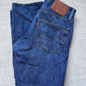 Levi's 501 blå jeans - Klassiska blå Levi's 501 jeans med raka ben och femficksdesign. Jeansen har knappgylf, bruna sömmar och läderpatch bak i midjan. Tillverkade i slitstarkt denim och passar perfekt till en avslappnad stil.