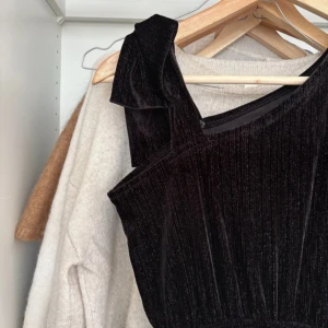 Svart sammetsjumper med oneshoulder från Zara - Säljer en lång svart sammetsjumper från Zara med oneshoulder och en ärm med lite silver glitter på. Klänningen har en snygg glansig yta och knappar i sidan. Perfekt för dig som vill sticka ut med en elegant look. Och en liten fläck som jag ska se går bort i tvätten💕 storlek 164 ifrån Zara!💞
