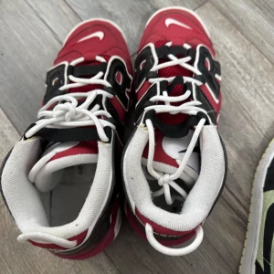 Nike Air More Uptempo röd/svart - Säljer ett par Nike Air More Uptempo sneakers i rött med stora svarta AIR-bokstäver på sidorna. Skorna har vit sula, vita snören och detaljer i svart och vitt. Perfekta för dig som gillar en sportig och retro look. Pris går att diskutera