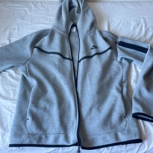 Grå zip-hoodie från Nike - Säljer en grå hoodie från Nike med hel dragkedja och svarta detaljer. Jackan har huva, långa ärmar och två fickor framtill. Perfekt för träning eller chill. Klassisk Nike-logga på bröstet.