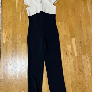 Svart och vit jumpsuit med kort ärm - Säljer en stilren jumpsuit i svart och vitt med korta ärmar och v-ringad detalj framtill. Jumpsuiten har en tvåfärgad design med vit överdel och svart nedtill, samt långa ben. Perfekt för dig som vill ha en chic och modern look.