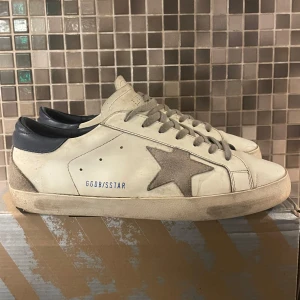 Vita sneakers från Golden Goose med grå stjärna - Säljer ett par vita sneakers från Golden Goose med grå stjärndetalj på sidan och mörkblå häl. Skorna har grå snörning och en platt sula. Säljer dessa för att jag har tröttnat och vill köpa nya. Det är en liten skada på under sidan men inget man ser eller tänker på och det kan fixas enkelt med lite lim