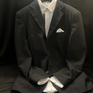 Svart kostym set med vit skjorta - Säljer en klassisk svart kostym med matchande svarta kostymbyxor och vit skjorta. Kavajen har två knappar och en bröstficka med vit näsduk. Perfekt för formella tillfällen.