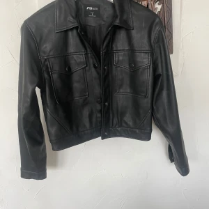 Svart skinnjacka från New Yorker - Säljer en svart skinnjacka från New Yorker i storlek S. Jackan har klassisk krage, två bröstfickor med lock och knappar framtill. Snygg och tidlös modell som passar till många olika stilar.