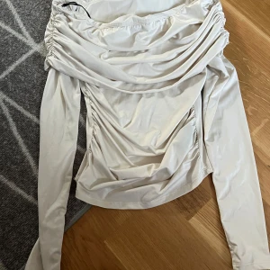 Beige offshoulder topp från Monki - Säljer en beige offshoulder topp från Monki med långa ärmar och snygga draperade detaljer framtill. Toppen har en figurnära passform och är tillverkad i ett mjukt, stretchigt material. Perfekt för att lyfta din outfit!