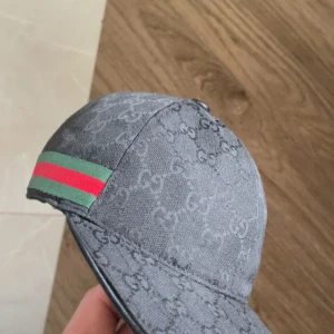 Gucci keps med monogram och röd-gröna ränder - Säljer en snygg svart keps från Gucci med klassiskt GG-monogram över hela kepsen och de ikoniska röd-gröna ränderna på sidorna. Kepsen har böjd skärm och justerbar rem baktill. Perfekt för dig som gillar exklusiva accessoarer.