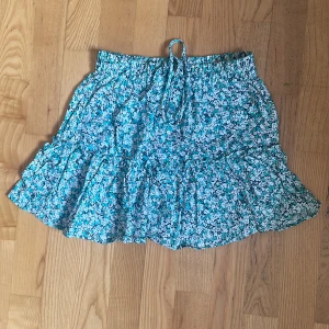 Blommig grön volangkjol från Zara - Supersöt kort kjol från Zara med grönt, svart och vitt blommönster och volangdetaljer. Resår i midjan med knytband för extra komfort. Perfekt för varma dagar och enkel att matcha med en enkel topp.