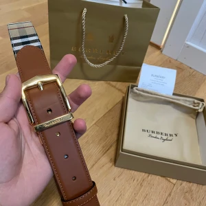Burberry bälte  - Säljer ett snyggt bälte från Burberry i brunt skinn med klassiskt beige, svart, vitt och rött rutigt tyg på ena sidan. Bältet har ett stort, guldigt spänne med Burberry-logga. Perfekt accessoar för att lyfta din outfit.