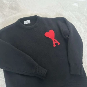 Svart stickad tröja från Ami med rött hjärta - Säljer en svart stickad tröja från Ami med ett stort rött hjärta och bokstaven A på bröstet. Tröjan har rund halsringning och långa ärmar med ribbade muddar. Perfekt för dig som gillar stilrena plagg med en twist.