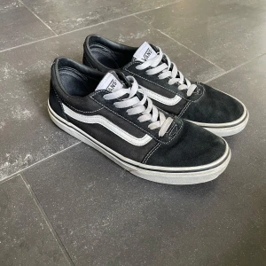 Svarta Vans Old Skool sneakers - Vans Old Skool sneakers i svart med vit sula och vit sidoremsa i storlek 39. Skorna har snörning och är tillverkade i en kombination av mocka och canvas. Den ena skon är lite skadad på sulan men det är inget som märks. 