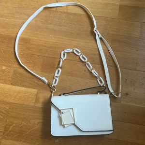 Vit axelväska med kedjedetalj - Snygg vit axelväska med justerbart axelband och dekorativ kedja i plast. Väskan har ett geometriskt spänne i guld och magnetlås. Insidan har ett fack med dragkedja. Perfekt för att lyfta din outfit!