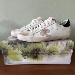 The Gallerist skor  - Snygga vita sneakers från The Gallerist med en unik klöverdetalj på sidan. Skorna har en beige mockadetalj på tån och svart häl. Perfekta för en stilren look. Stl 42. 