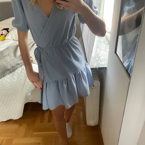 Ljusblå omlottklänning med puffärm - Supersöt ljusblå omlottklänning med puffärmar och knyt i midjan. Klänningen har volang nertill och är kort i modellen. Perfekt för vår och sommar💕🌸