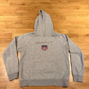 Grå hoodie från GANT original shield logga  - Säljer en klassisk grå hoodie från GANT med broderad logga och sköld på bröstet. Tröjan har huva, magficka och ribbade muddar vid ärmslut och nederkant. Perfekt för en avslappnad stil. Storleken är M, men passar även S. Helt oanvänd. Nypris 1 000, pris kan diskuteras vid snabb affär 