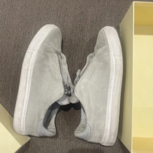 Axel arigatos skor - Säljer ett par ljusgråa sneakers i mocka med dragkedja framtill istället för klassisk snörning. Skorna har en platt vit sula och rund tå, vilket ger en stilren och modern look.