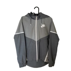 Grå vindjacka från Nike - Snygg grå vindjacka från Nike med huva och vit dragkedja. Jackan har två olika grå nyanser, vita detaljer och Nike-logga på bröstet. Justerbar huva med snören och elastiska muddar vid ärmslut. Perfekt för blåsiga dagar. Knappt använd, sitter som S typ. skrvw för mer information 😇😇😇
