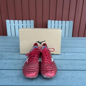 Röda Mizuno Morelia Neo fotbollsskor - Säljer ett par röda Mizuno Morelia Neo fotbollsskor med vita detaljer och snörning. Skorna har rund tå och platt sula med dobbar, perfekt för spel på både konst- och naturgräs. Klassisk design med Mizuno-logga på sidan. Välanvända, skick enligt bilder. 