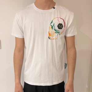 Vit t-shirt med broderad döskalle från Philipp Plein - Säljer en vit t-shirt från Philipp Plein med en färgglad broderad döskalle och PP-logga på bröstet. T-shirten har normal passform och är tillverkad i mjuk bomull. Perfekt för dig som vill sticka ut med en unik detalj.