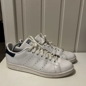 Adidas Stan Smith - Klassiska Adidas Stan Smith sneakers i vitt skinn med mörkblå detalj på hälen. Skorna har snörning, perforerade ränder på sidorna och rund tå. Perfekta för en clean och tidlös stil. Ikonisk logga på plösen och hälen.