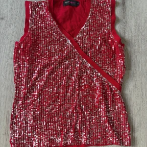 Röd paljettopp från MariPhilippe - Röd ärmlös topp från MariPhilippe med v-ringning och omlottkänsla framtill. Hela framsidan är täckt av glittrande paljetter som ger en riktigt festlig vibe. Baksidan är i mjuk bomull utan paljetter. Perfekt för fest!