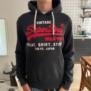 Svart Superdry hoodie  - Svart hoodie från Superdry med stort rött och vitt tryck på bröstet. Klassisk känguruficka, vita snören i huvan och ribbade muddar. Mjuk bomullskänsla och avslappnad passform, perfekt för chill dagar.