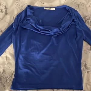Blå långärmad topp från NA-KD - Säljer en snygg blå långärmad topp från NA-KD med draperad krage. Toppens material är glansigt och stretchigt, vilket ger en cool och modern look. Perfekt för dig som vill sticka ut med en färgstark och trendig stil.