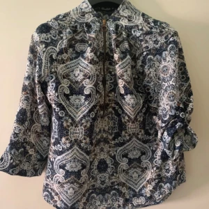 Mönstrad skjorta från Miss Millo M - Snygg skjorta från Miss Millo i storlek M med unikt paisleymönster i blått, beige och vitt. Skjortan har trekvartsärmar med knyt vid ärmslut och en cool dragkedja framtill med ringdetalj. Perfekt för dig som vill sticka ut med din stil.